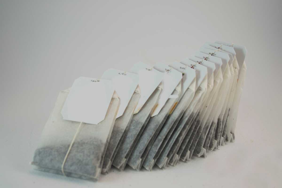 A neat row of teabags is displayed, all leaning against each other. Each teabag has a white label attached with string, and the contents of the bags appear to be black tea. 