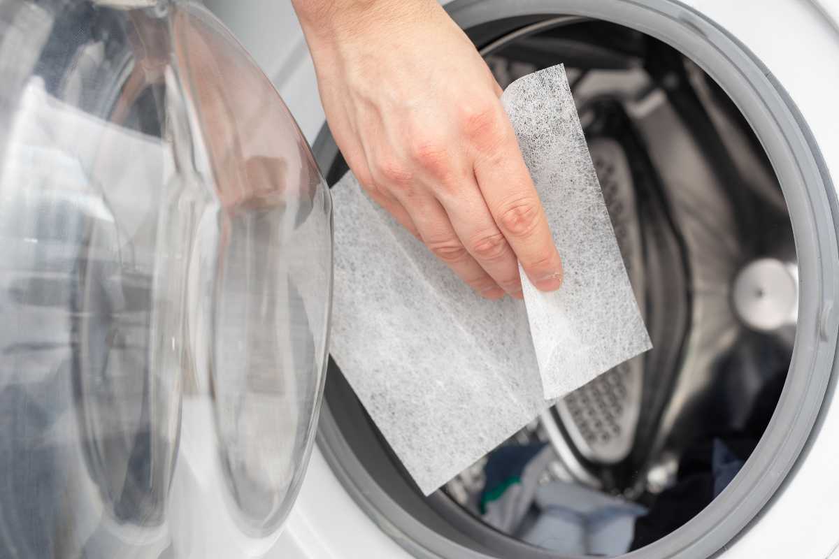 A hand placing a dryer sheet inside an open front-loading washing machine. 