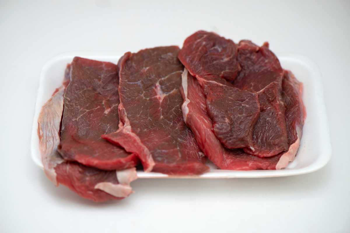 A white styrofoam tray contains several slices of raw red meat, likely beef. The meat is marbled with thin veins of fat and appears fresh.