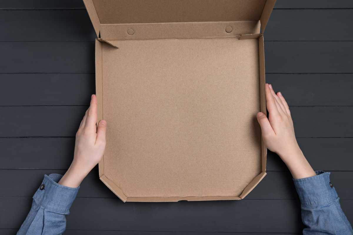 Two hands holding an open, empty cardboard pizza box on a dark, textured background. The person is wearing a blue long-sleeved shirt.