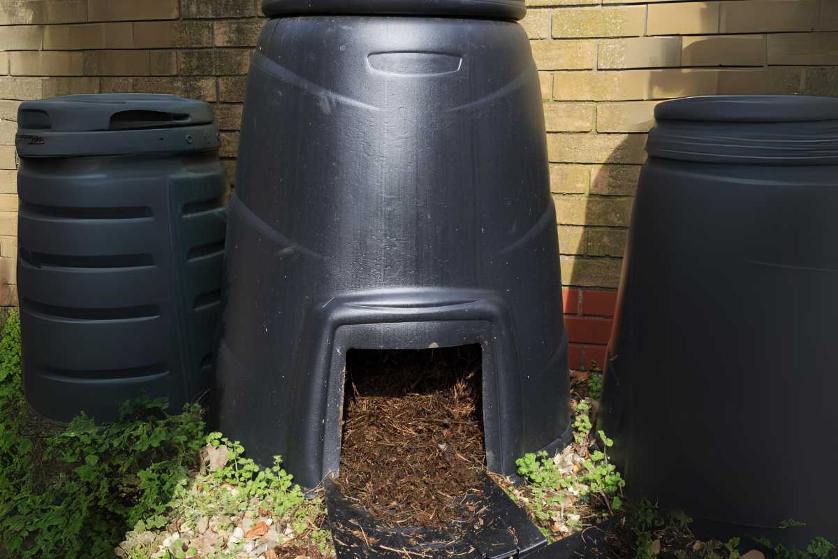 Three large black plastic compost bins are set against a brick wall, with one in the center having an open compartment at the bottom showing composting material. 