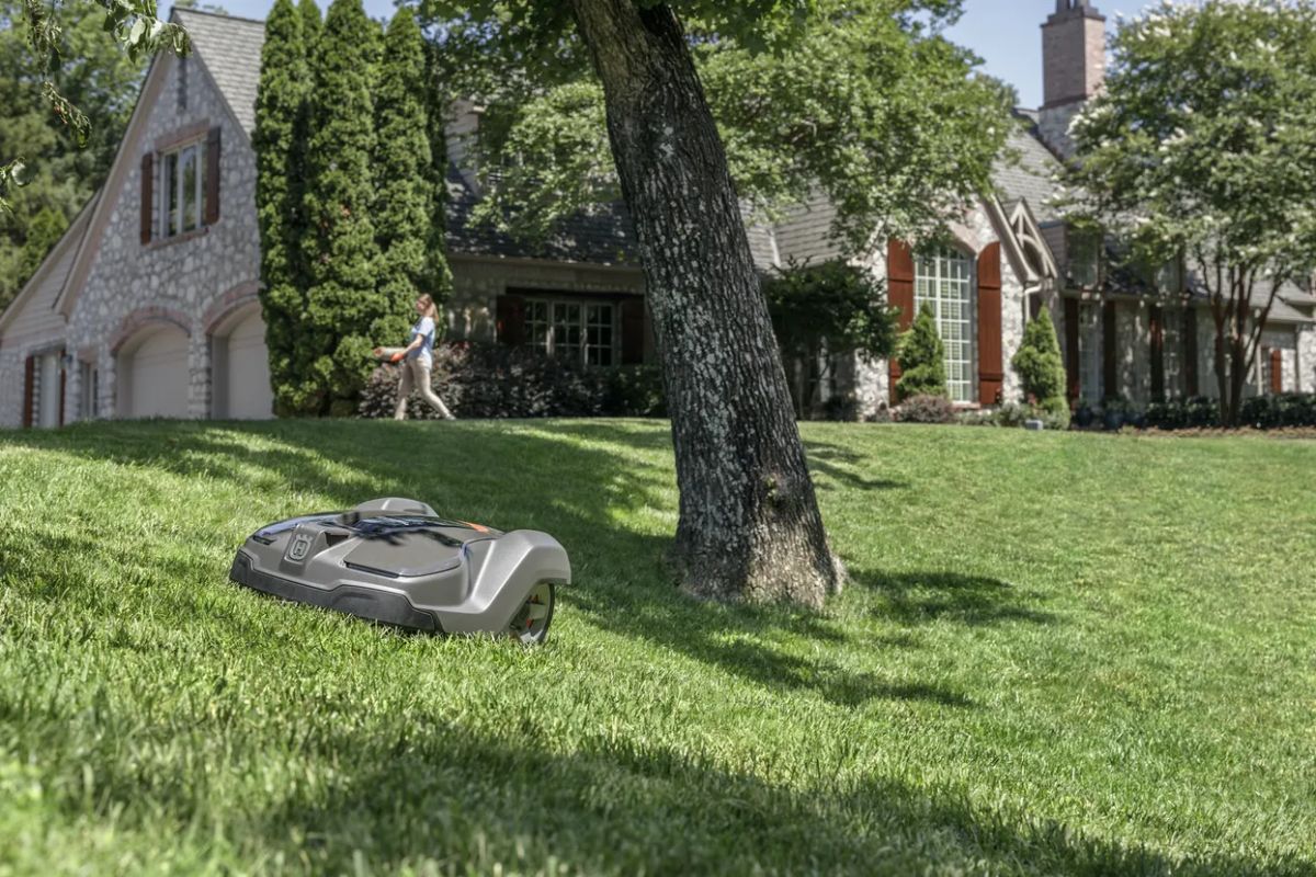 A Husqvarna Automower 430X is cutting grass on a sloped lawn in front of a large house surrounded by trees. 