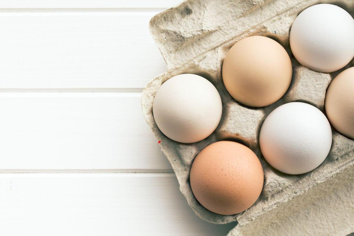 A close-up of a half-dozen eggs in a cardboard carton on a white wooden surface. 