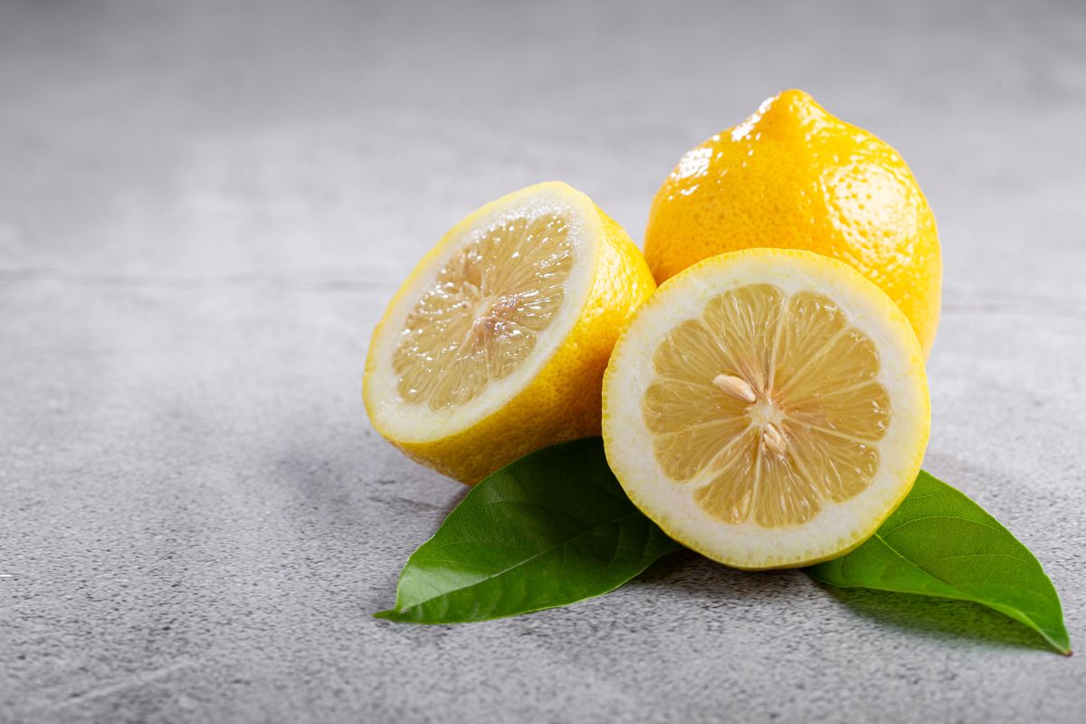 Two lemon halves and a whole lemon are placed on a gray surface with two green leaves underneath. 