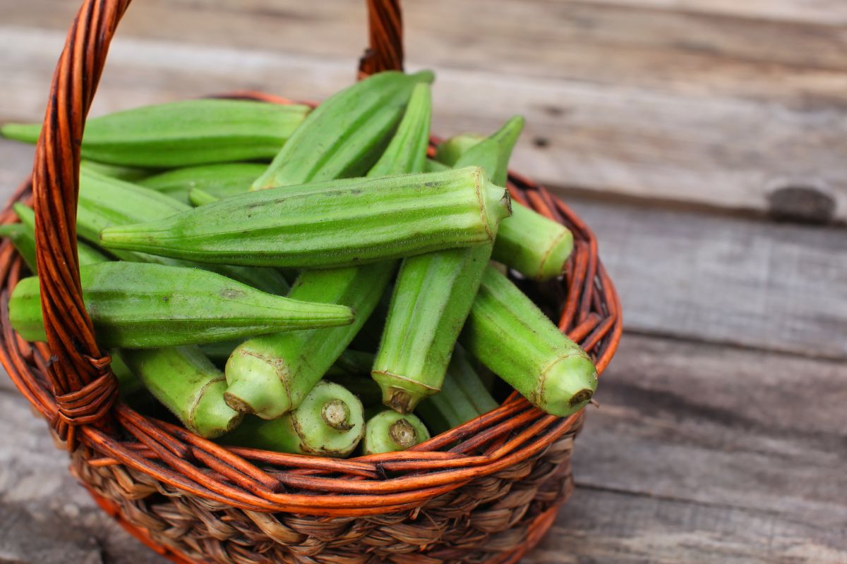 A woven basket filled with fresh green okra, placed on a rustic wooden surface. 