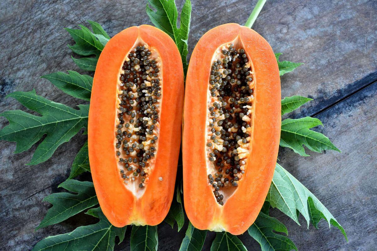 Sliced papaya with black seeds is displayed on a bed of green papaya leaves against a rustic wooden background.