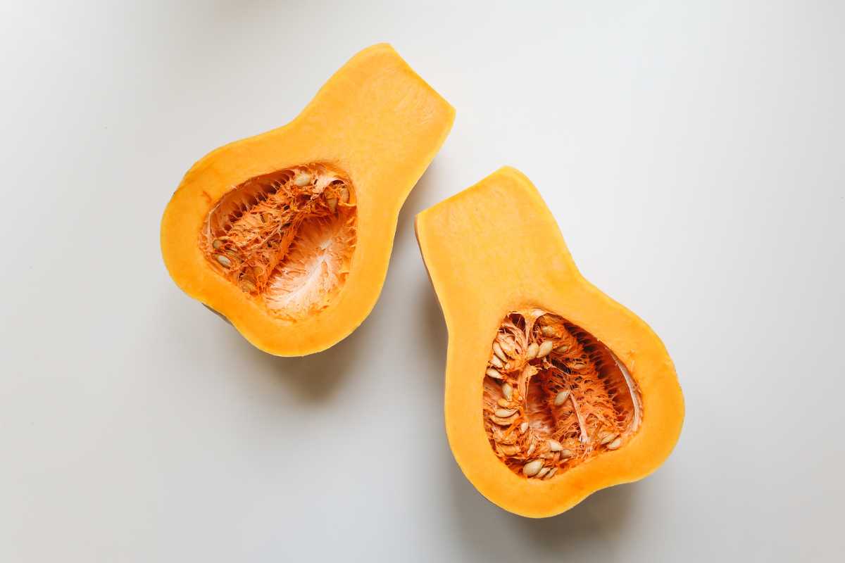 Two halves of a butternut squash lie on a light-colored surface. The squash has a smooth, orange-yellow outer skin with firm flesh of a similar color and a hollow cavity containing seeds and fibrous strands. 