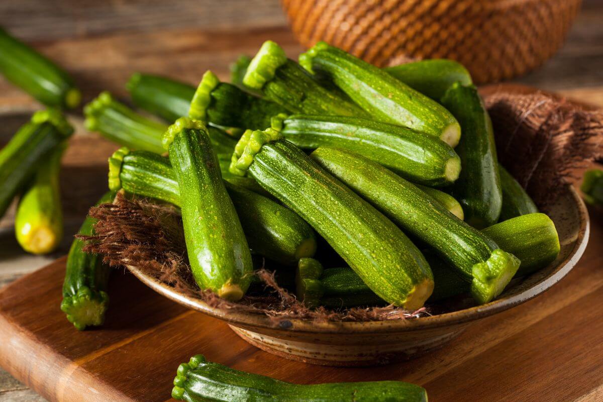 A ceramic bowl filled with fresh green zucchinis sits on a wooden surface.