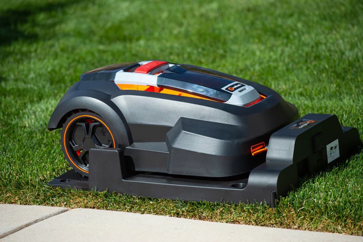 A sleek, black mowro rm24 robotic lawn mower sits on a neatly trimmed grassy lawn, with red and orange accents. The machine is docked on its charging base, located on the grass near a paved sidewalk.