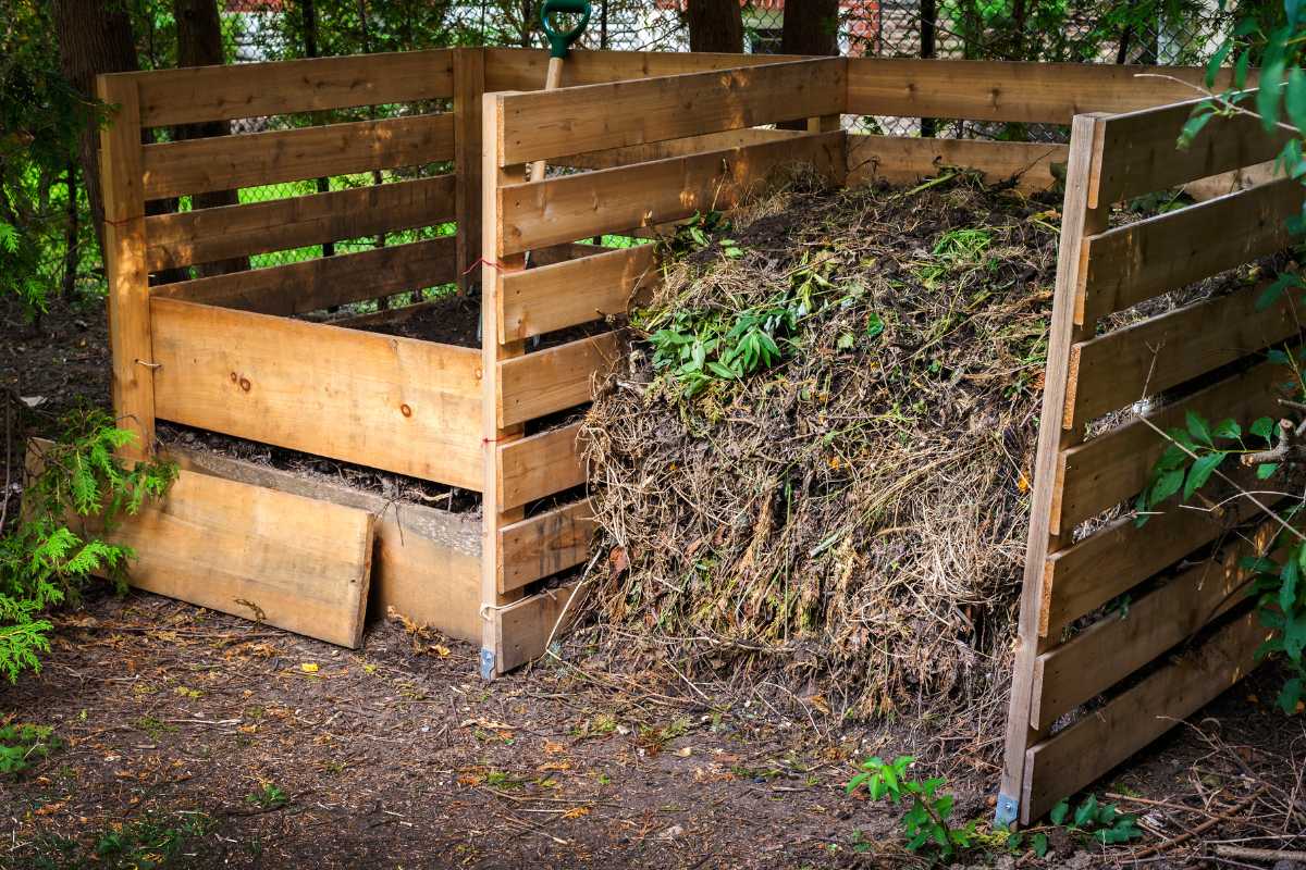 An open-air wooden compost bin filled with decomposing leaves, grass, and other organic matter is situated outdoors. 