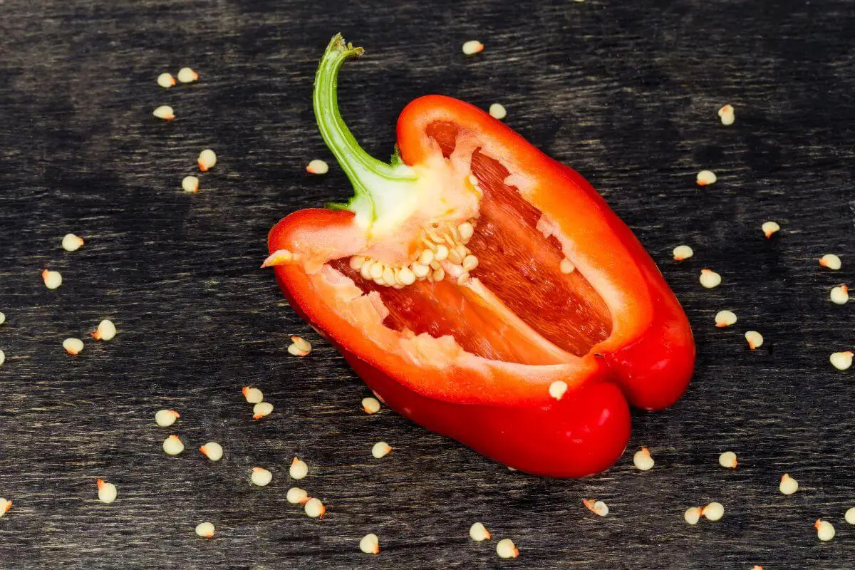 A halved red bell pepper lies on a dark wooden surface with seeds scattered around it. The interior of the bell pepper, including its stem and seeds, is visible, showcasing its vibrant red hue and texture.