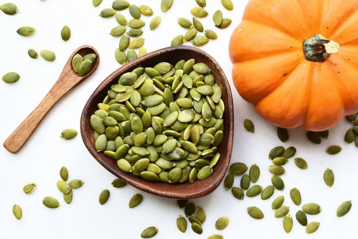 A wooden bowl filled with green pumpkin seeds sits beside an orange pumpkin. A wooden spoon with a few seeds on it is visible, and scattered seeds are spread on a white surface.