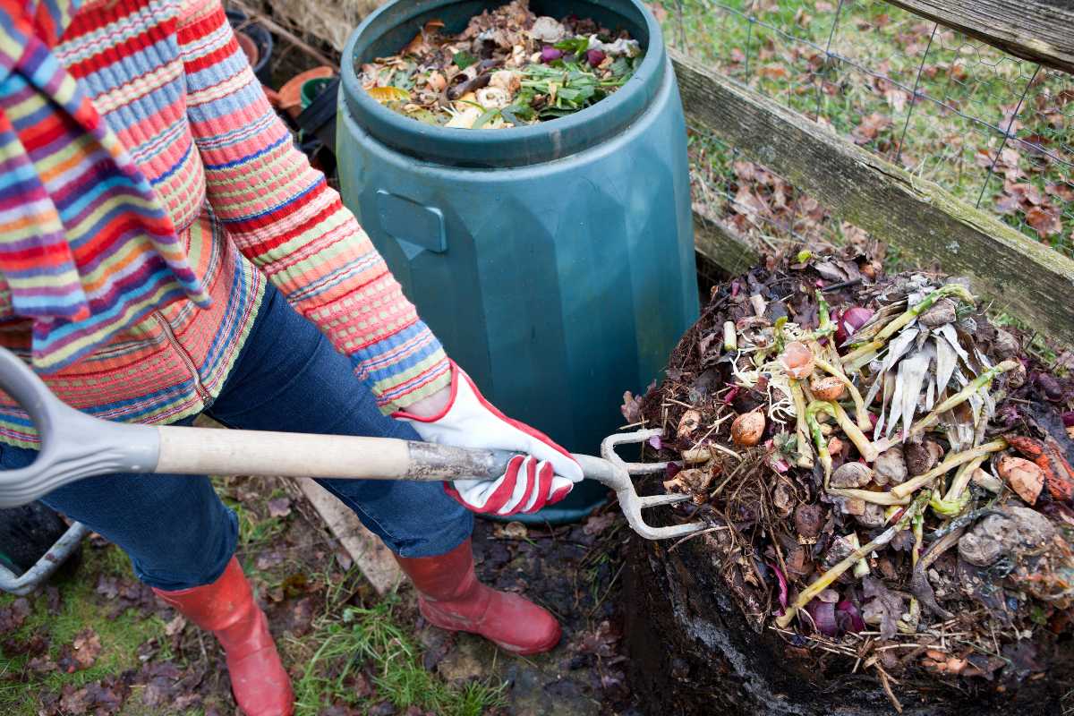 A person wearing a colorful striped sweater, red gloves, and red boots uses a pitchfork to turn over compost in an outdoor bin. 