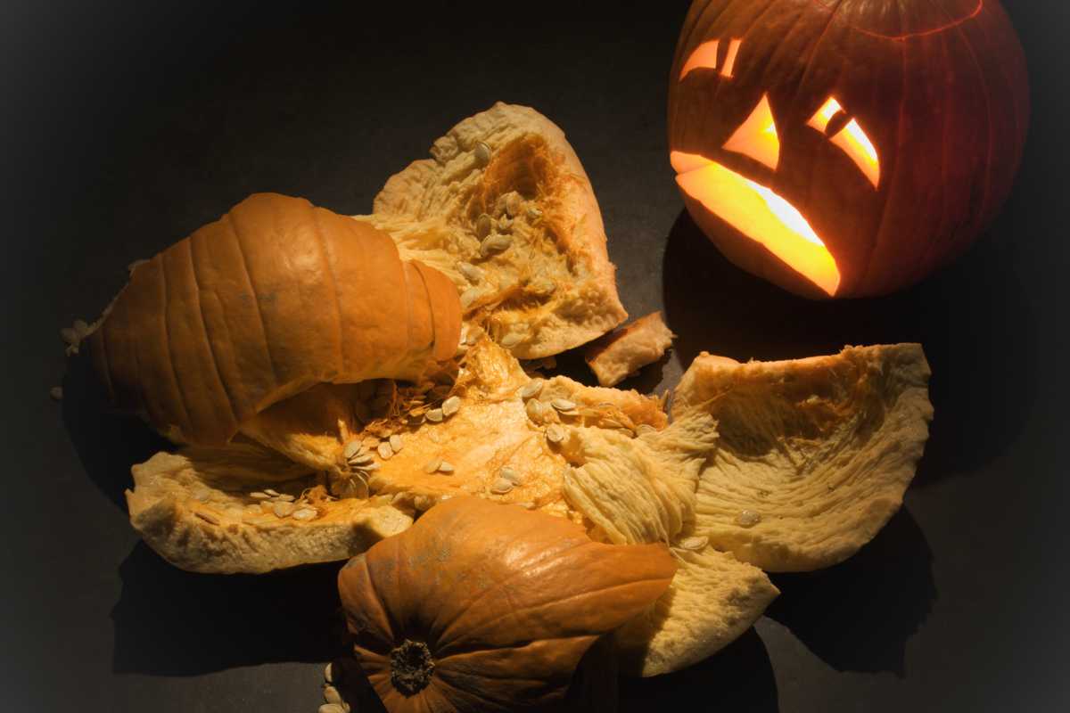 A lit jack-o'-lantern sits on a dark surface next to several pieces of broken, uncarved pumpkin flesh and seeds.