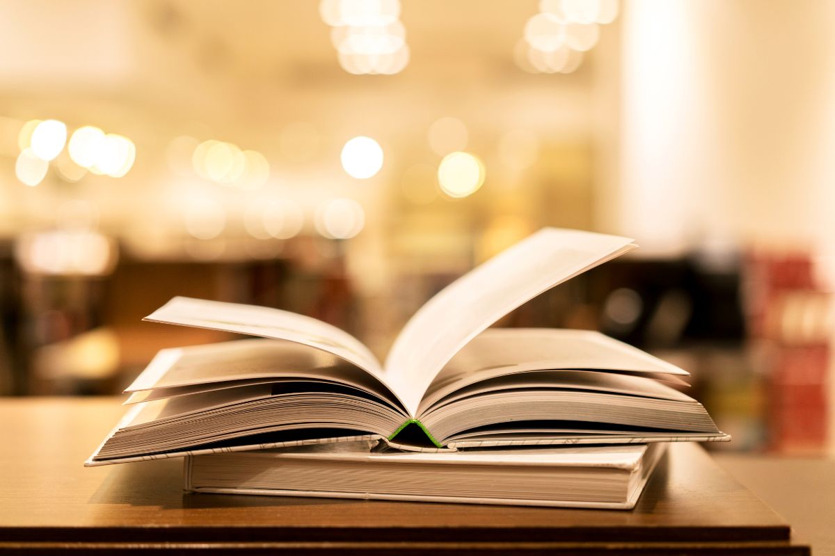 An open book resting on top of a closed book on a wooden table in a warmly lit room.