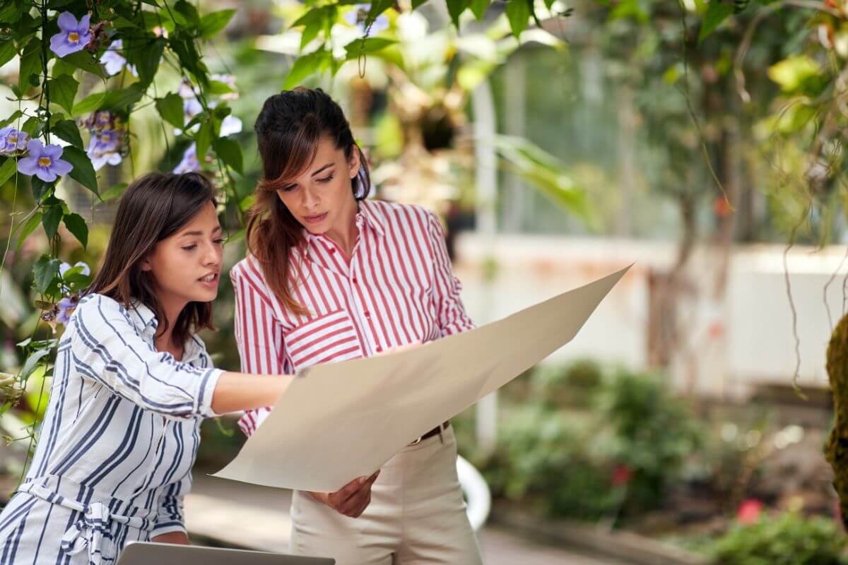 Two women in striped shirts, one white and one red, stand in a garden planning while looking at a large sheet of paper. 