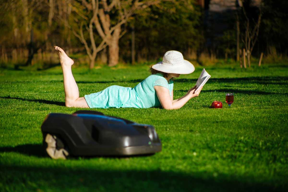 A person wearing a large sunhat and light green dress is lying on the grass on their stomach, reading a book. 