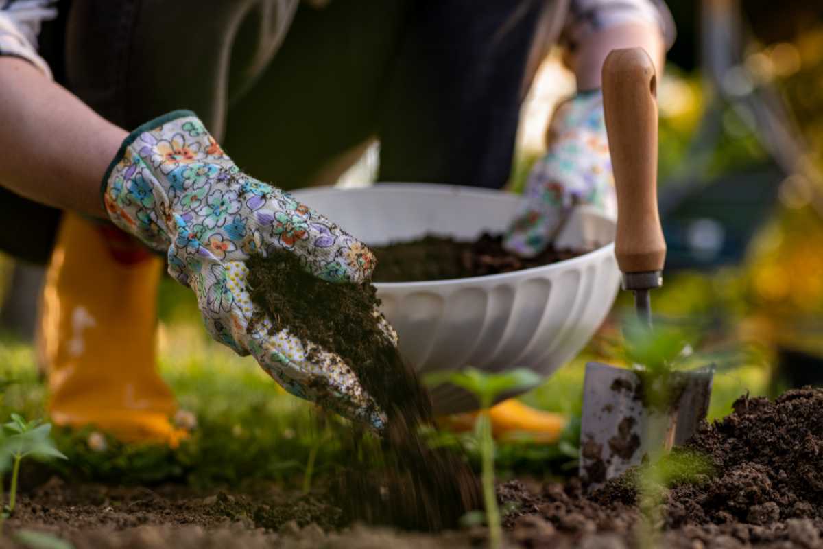 A person wearing colorful floral gardening gloves and yellow boots is scooping organic soil amendments from a white bowl with one hand while holding a small gardening trowel in the other.