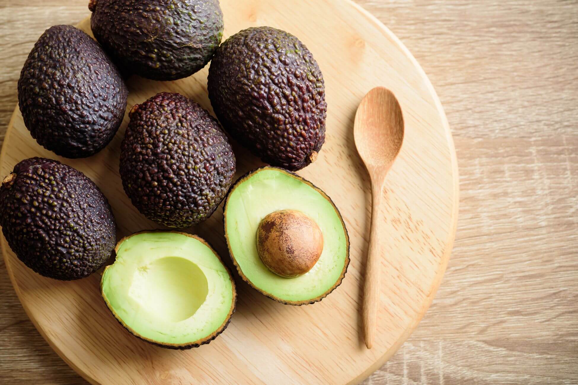 A wooden round cutting board with six avocados, including one halved to show its seed and green flesh.