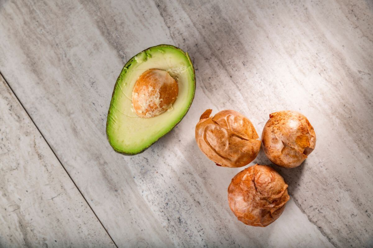 A halved hydroponic avocado with the seed exposed lies on a light wooden surface next to three round, golden-brown seeds.