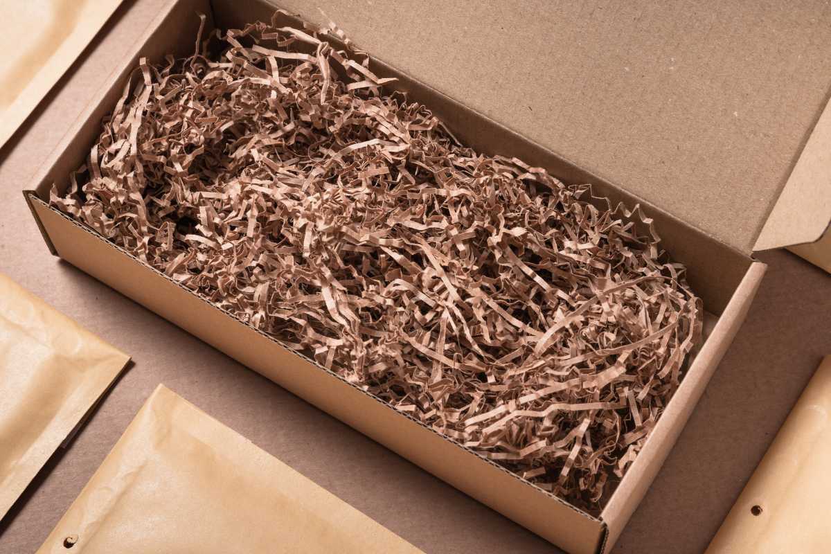 An open cardboard box filled with shredded cardboard paper is placed on a flat surface surrounded by several closed brown paper envelopes.