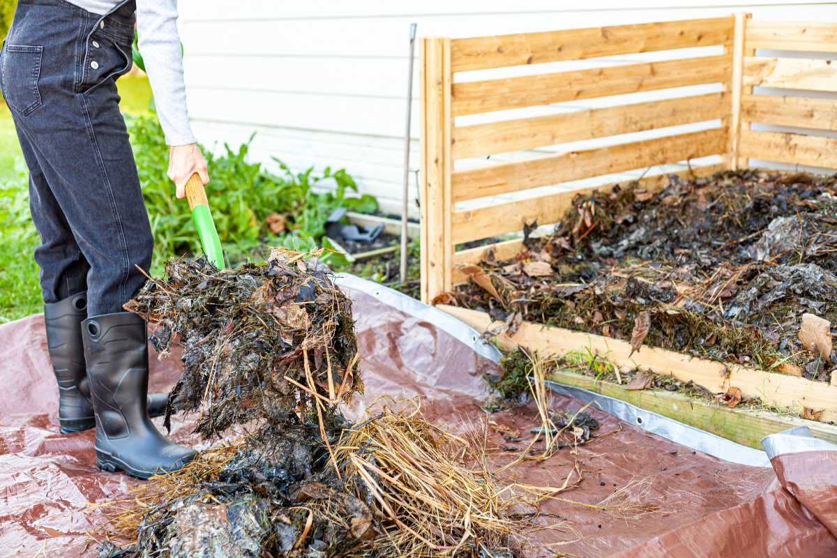 A person dressed in dark pants, long sleeves, and rubber boots uses a green garden fork to turn compost in a wooden bin filled with leaves, soil, and organic material. 