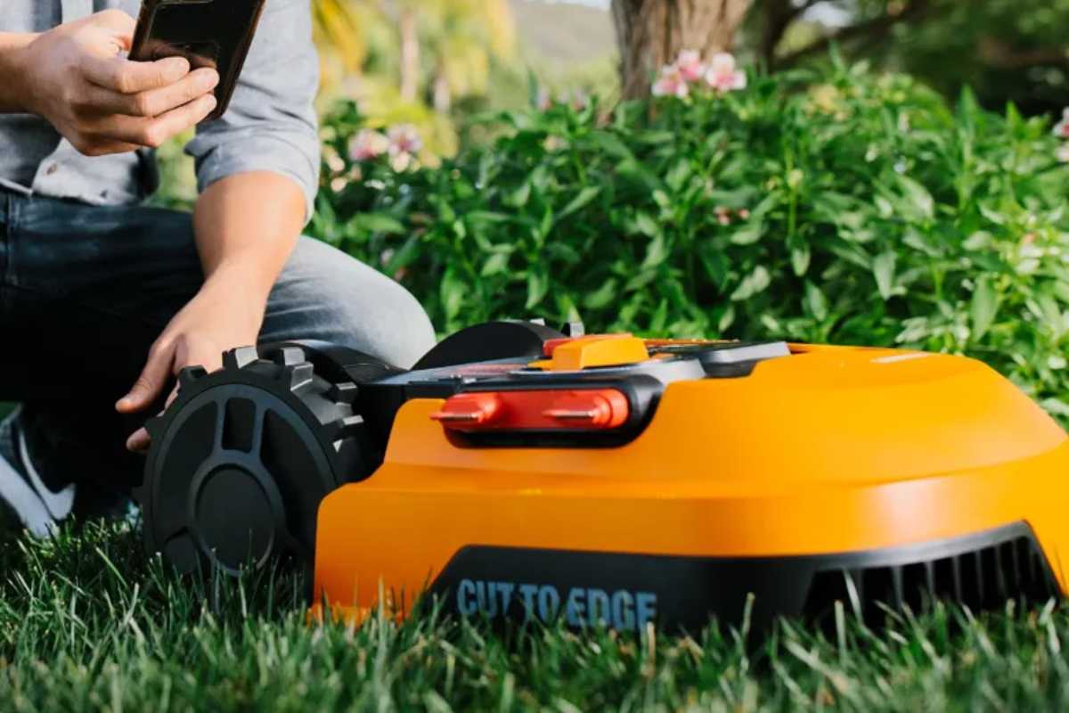 A person is crouching on grass, holding a smartphone next to an orange Worx WR150 robotic lawn mower.