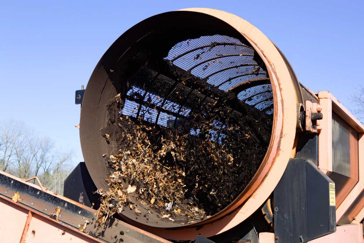 A large compost machine's drum with a mesh screen rotates, sifting out leaves and small debris for composting.