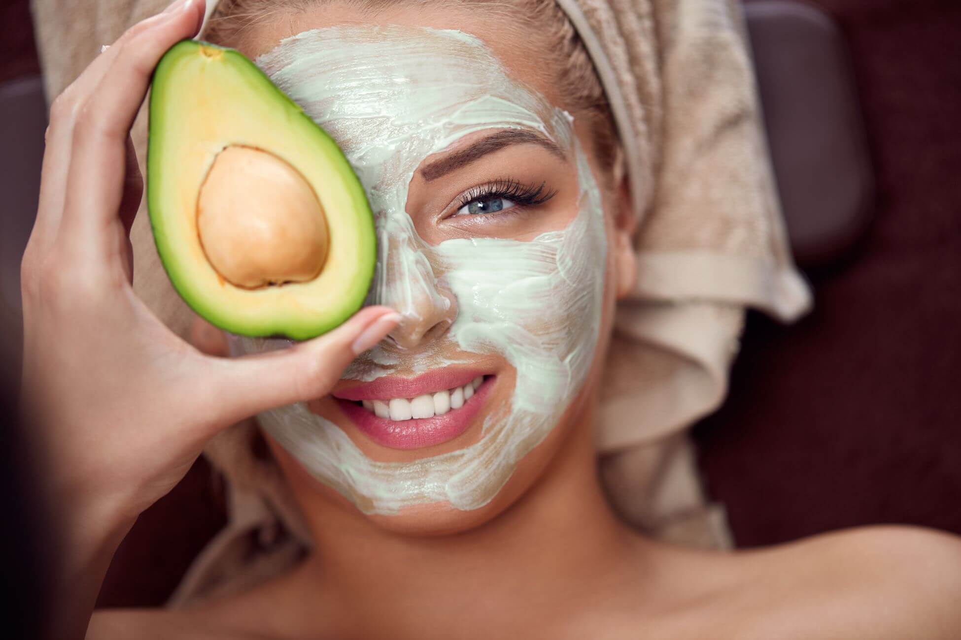 A smiling woman with a towel wrapped around her head applies a creamy avocado face mask. 
