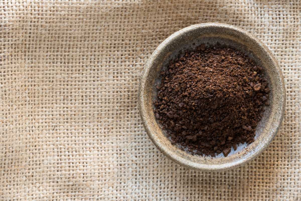 A small stone bowl filled with ground coffee sits on a beige burlap surface.