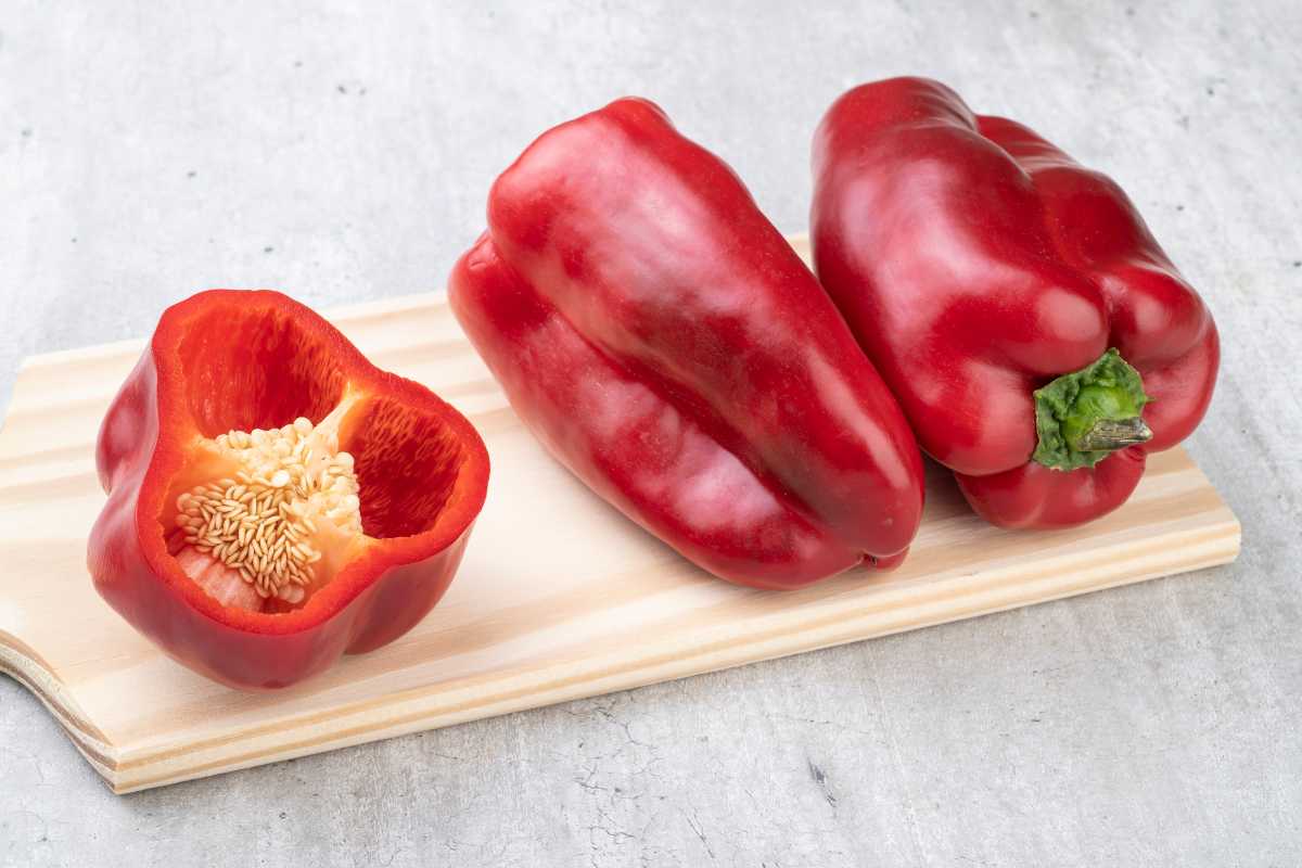 Three red bell peppers are on display: one cut in half with seeds visible, and two whole. They are laid on a light-colored wooden cutting board against a gray background.