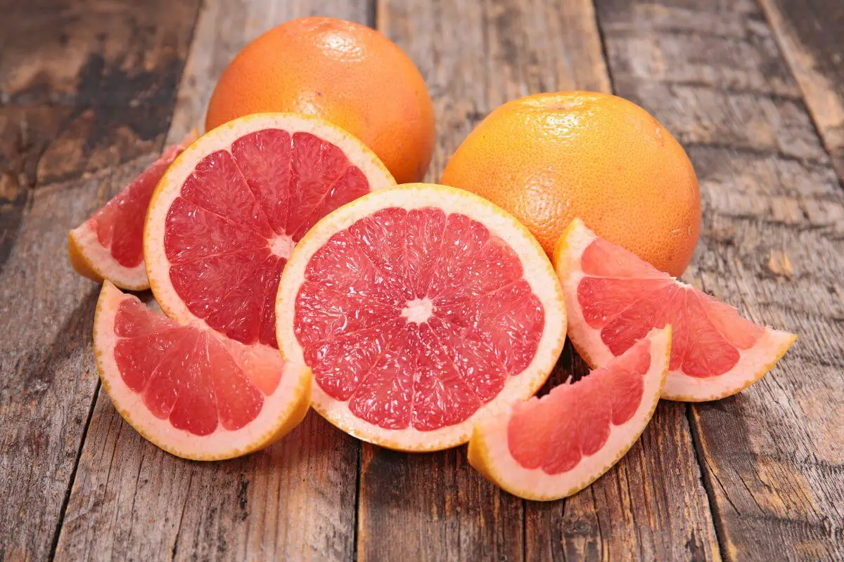 Several halved and sliced grapefruits placed on a rustic wooden surface. The grapefruit flesh is bright pink and juicy, contrasting with the yellowish-orange rind. 