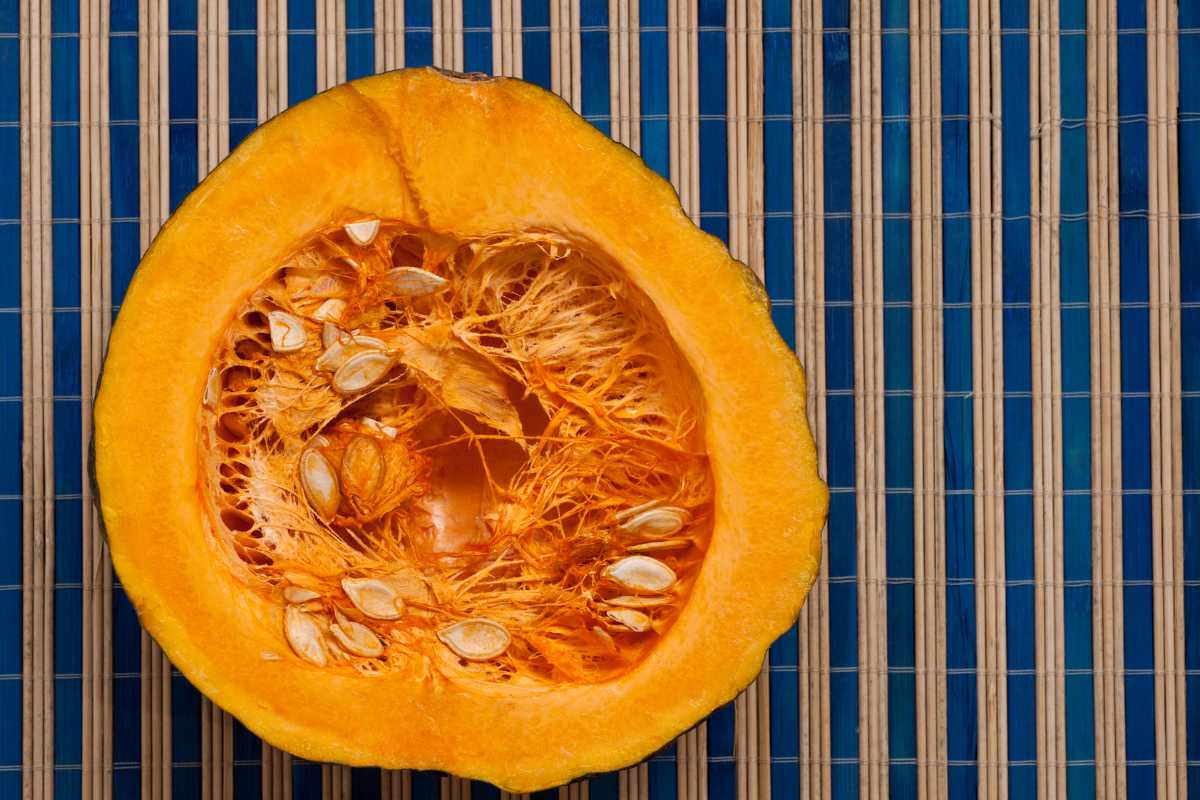 A halved pumpkin laid on a blue-striped bamboo mat. The pumpkin's flesh is vibrant orange with seeds and fibrous strands visible.