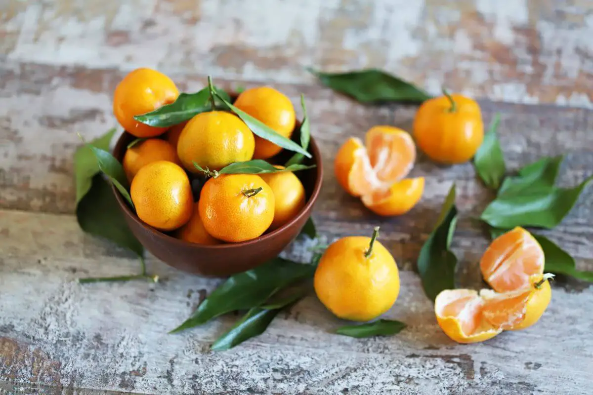A bowl filled with fresh, whole mandarins with their green leaves still attached sits on a rustic wooden surface. 