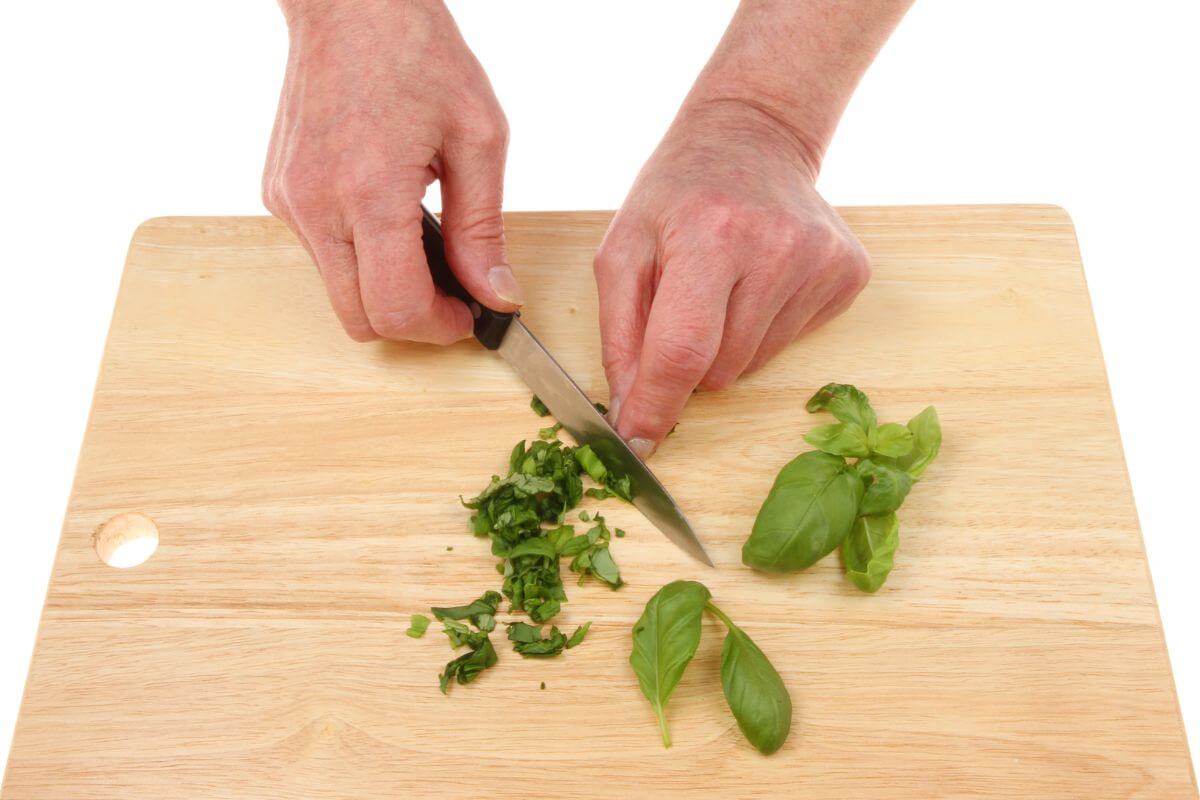 A pair of hands chopping fresh basil leaves on a light wooden cutting board with a knife.