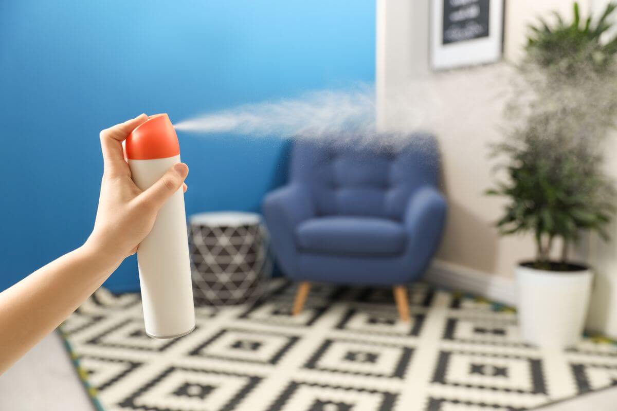 A person sprays aerosol air freshener from a white can with an orange nozzle in a living room featuring an air purifying plant, geometric black and white rug, blue armchair, and potted plant. 