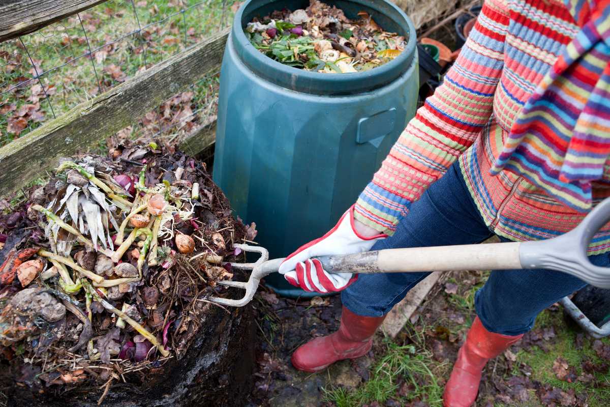 A person wearing a colorful striped sweater and red boots is turning a compost pile with a garden fork. 