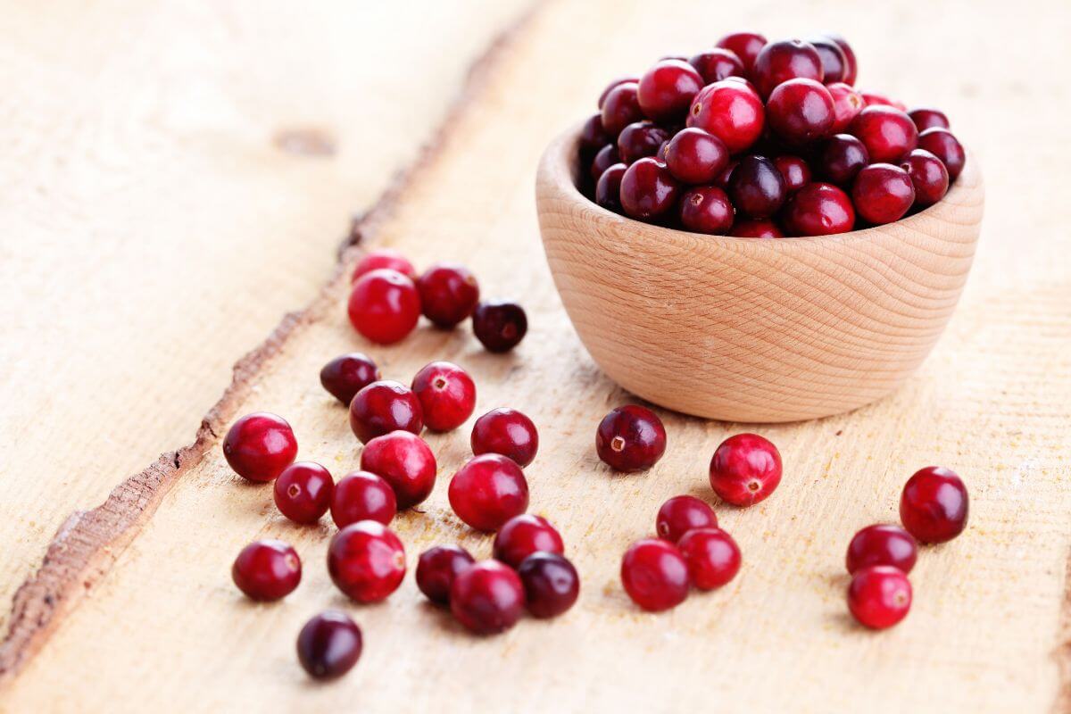 A small wooden bowl filled with ripe cranberries sits on a light wooden surface.