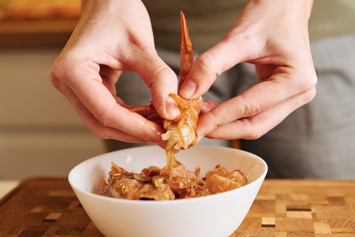 A pair of hands is peeling shrimp over a white bowl, removing the shell from a cooked shrimp. 