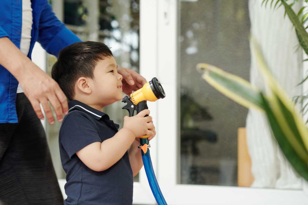 A child wearing a dark shirt holds a garden hose with a yellow nozzle, aiming it at the spider plants. 