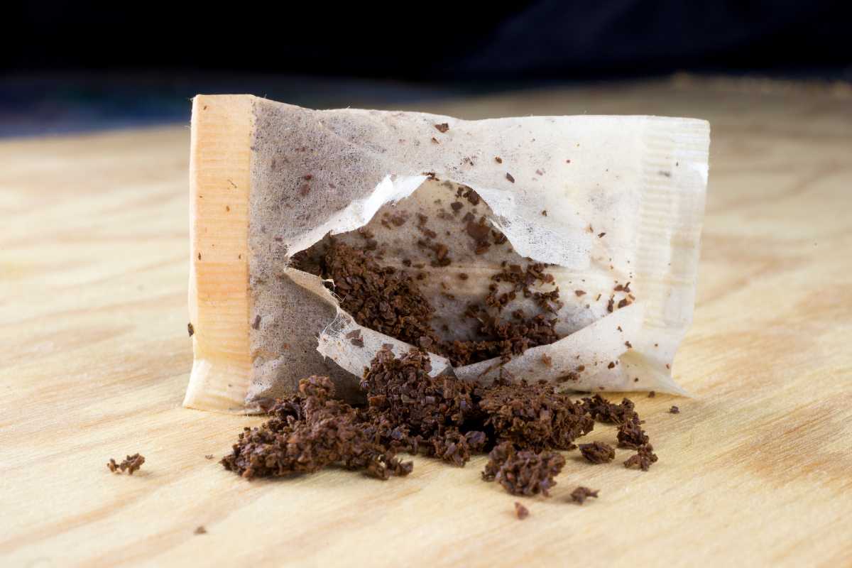 An opened tea bag lies on a wooden surface with loose tea leaves spilled out from the tear. 