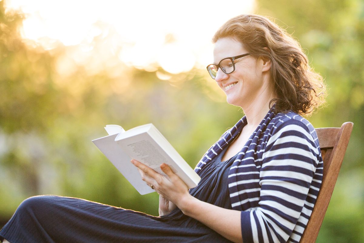 A woman with curly hair and glasses sits on a wooden chair outdoors, smiling as she reads a book about bonsai.