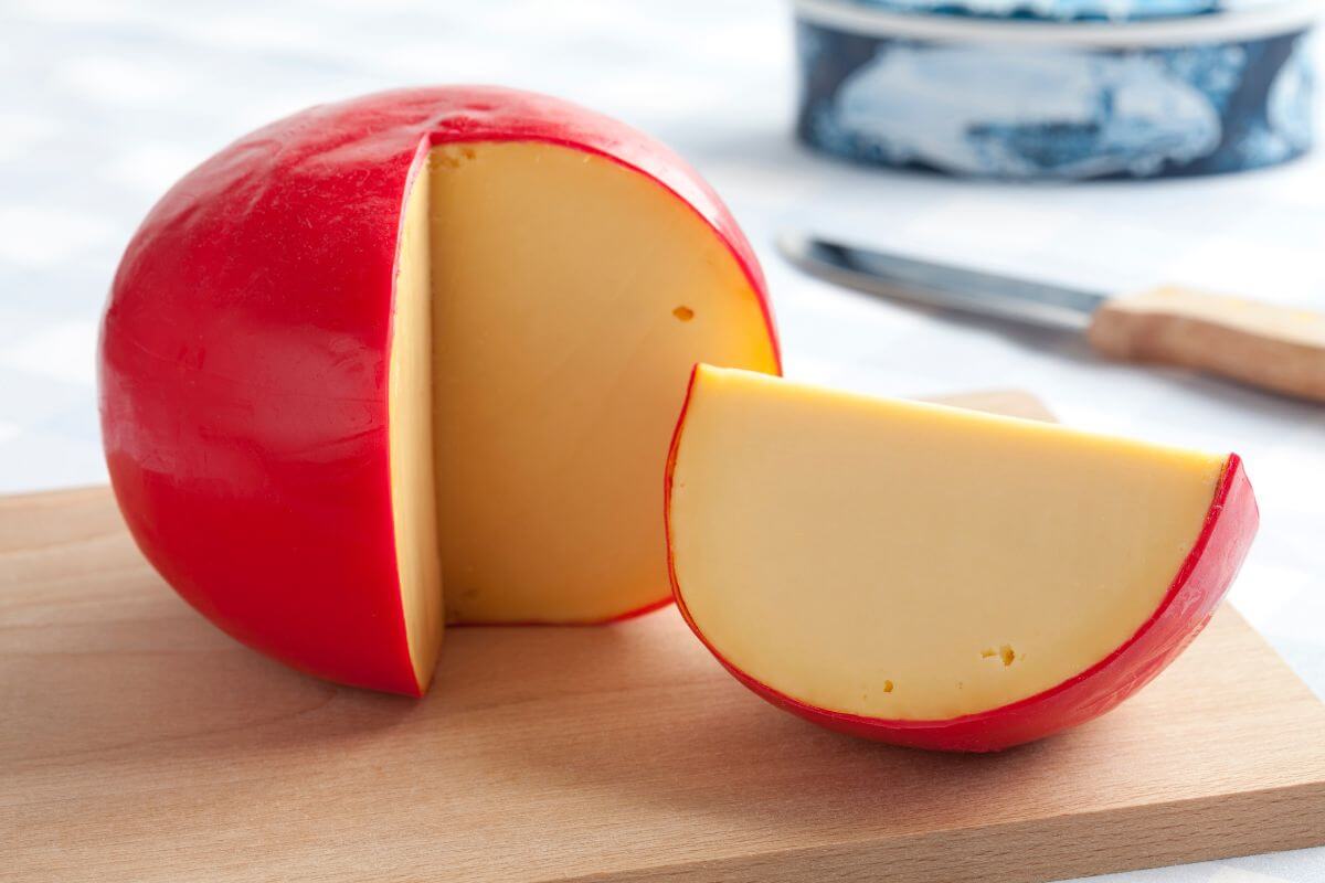 A round wheel of Gouda cheese with a red wax coating is shown on a wooden cutting board.