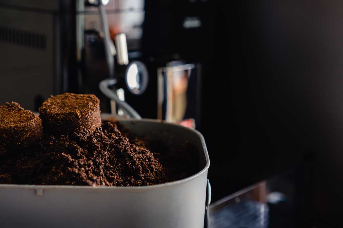 Coffee grounds in a container, with a coffee machine blurred in the background. The dark brown, densely packed grounds suggest they are freshly used and ideal for compost. 