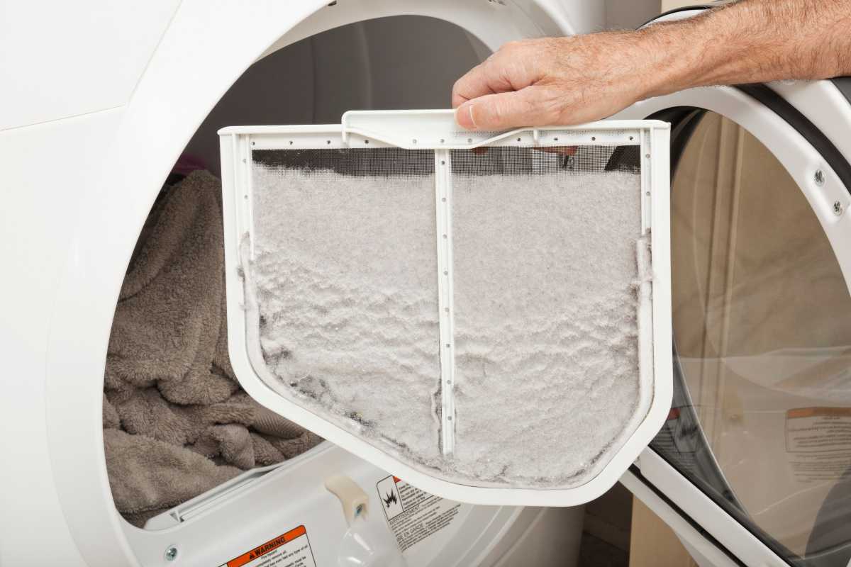 A person is holding a dryer lint filter filled with lint. The filter is being removed from a front-loading dryer, which is open and contains an assortment of fabric. 