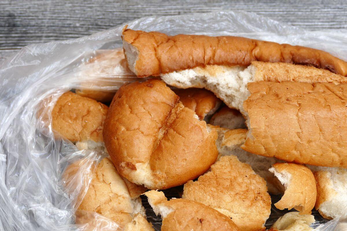 A collection of broken pieces of stale bread rolls, ideal for compost, are spread out in a partially transparent plastic bag on a wooden surface.