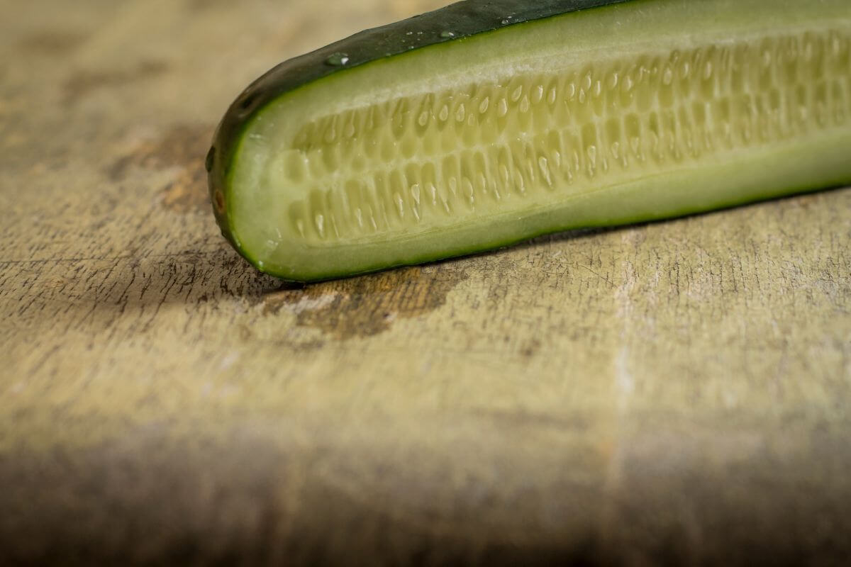 Close-up of a sliced cucumber laying on a textured wooden surface. The cucumber's interior seeds and fresh, juicy flesh are visible, and the outer green skin shows a slight curve. 