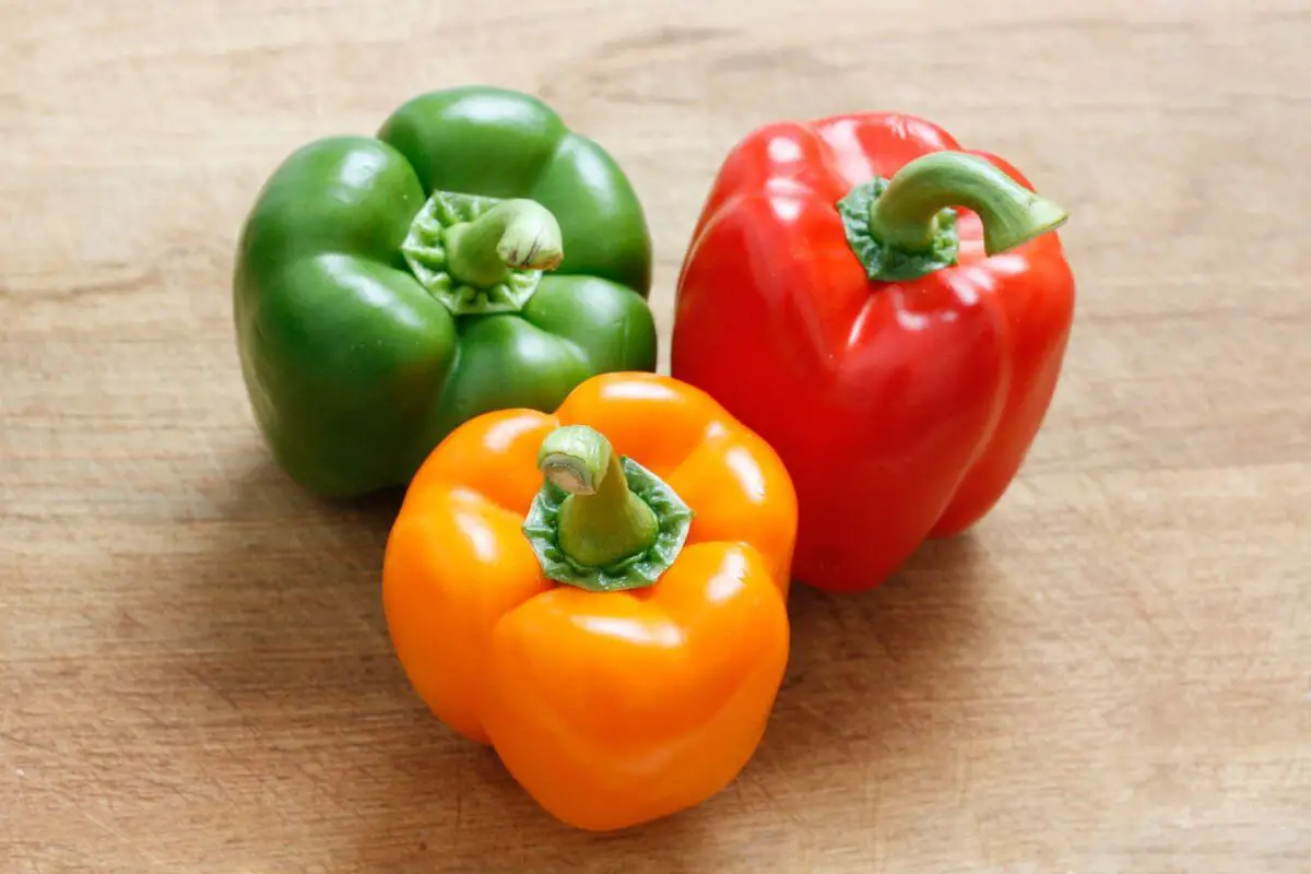 Three bell peppers are shown on a wooden surface. One is green, another is red, and the third is yellow-orange. They are fresh with their stems attached. 