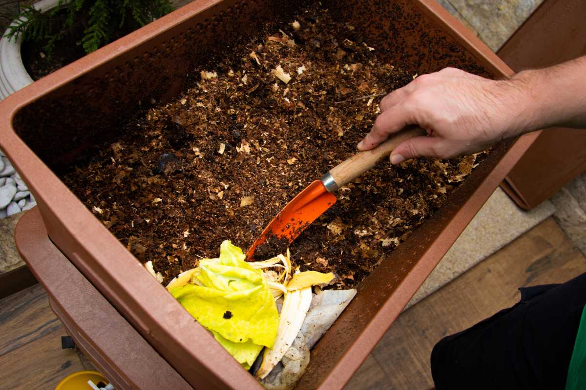 A person using a small gardening shovel to mix decomposing organic matter, including yellow vegetable peels, in a brown compost bin. 