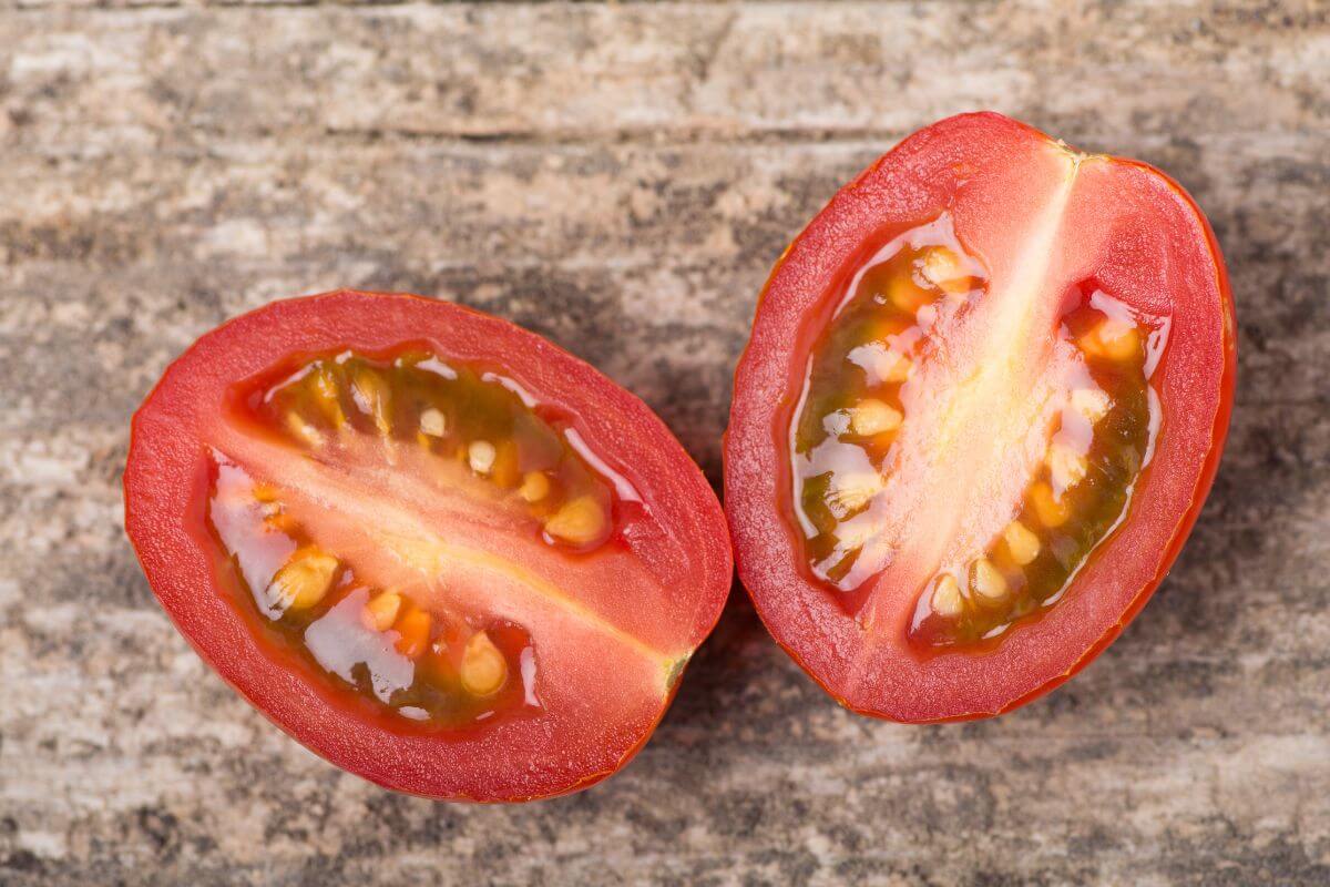 A tomato sliced in half lengthwise, resting on a rustic wooden surface. The tomato's vibrant red flesh is visible.